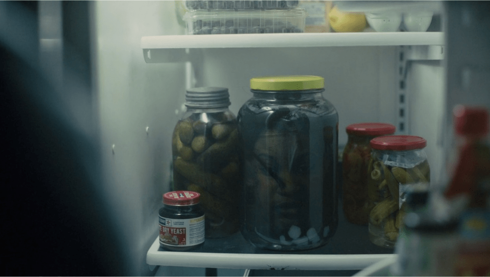 An opened refrigerator showing bottles with a pickle jar containing a head